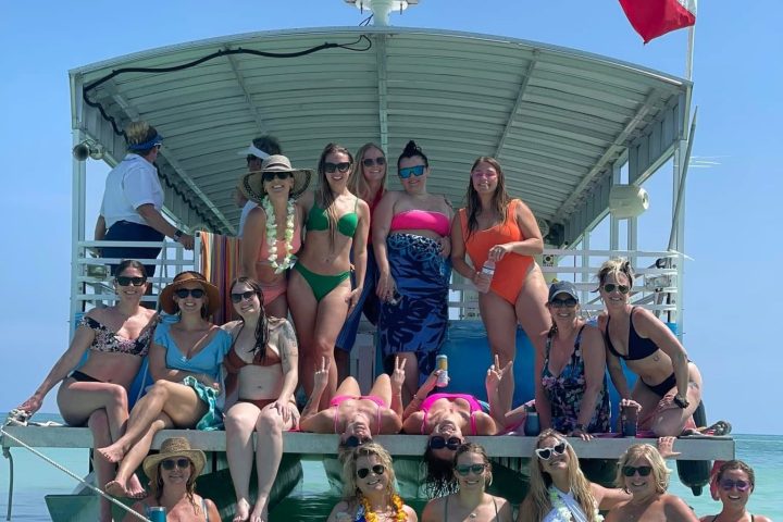 Group of women in swimsuits posing on a boat in clear water under a blue sky.
