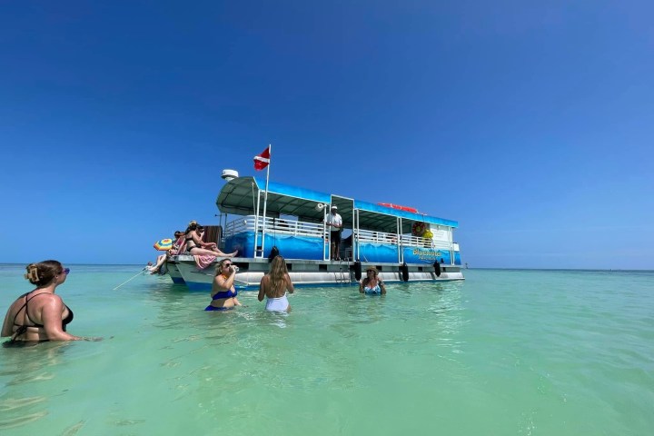 People swimming near a pontoon boat in clear turquoise water under a blue sky.