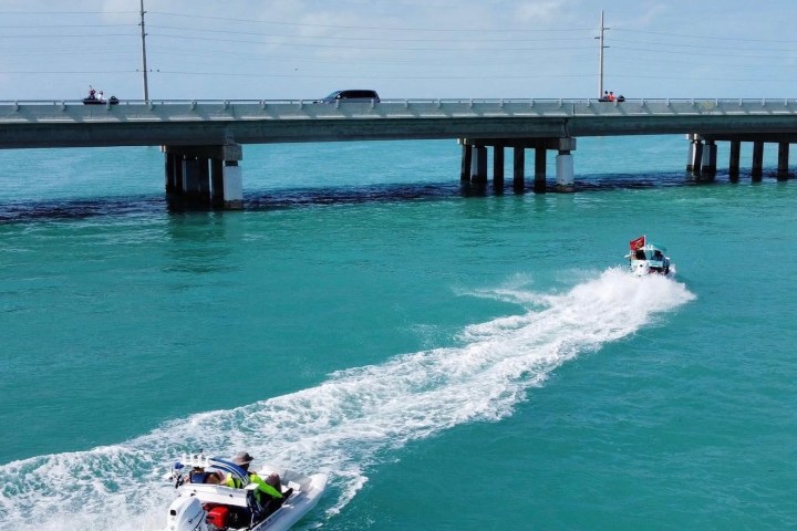 Two motorboats speed under a bridge over turquoise ocean water on a sunny day.