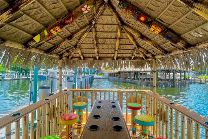 Tiki hut with colorful stools over water, surrounded by bamboo railing.