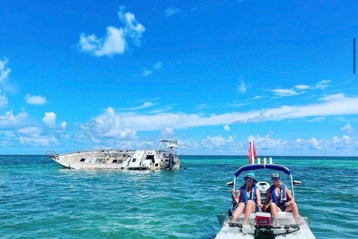 Two people on a motorboat near a shipwreck in clear turquoise water under a blue sky.