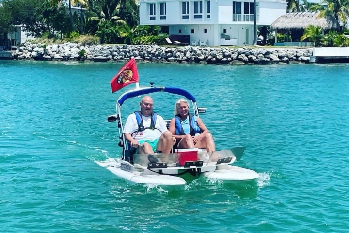 Two people on a small pedal boat in tropical waters, with houses and palm trees in the background.
