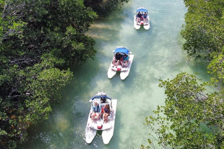 Three small boats with people navigate through a narrow, tree-lined waterway.