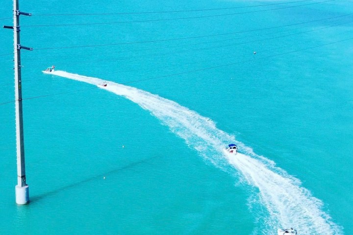 Boats creating wakes on turquoise water near a power line pole and small island in the distance.